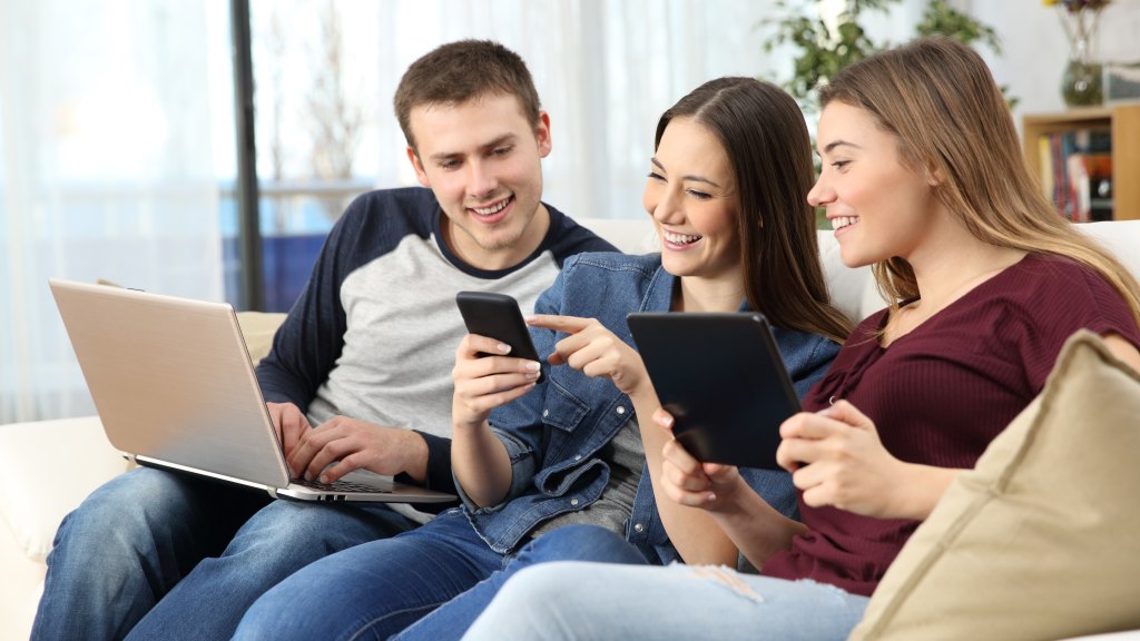 Três pessoas sentadas no sofá usando dispositivos conectados à rede Wi-Fi 6: um homem com notebook, uma mulher no meio com celular e outra mulher com tablet. Todos sorriem, aproveitando as vantagens do Wi-Fi 6.