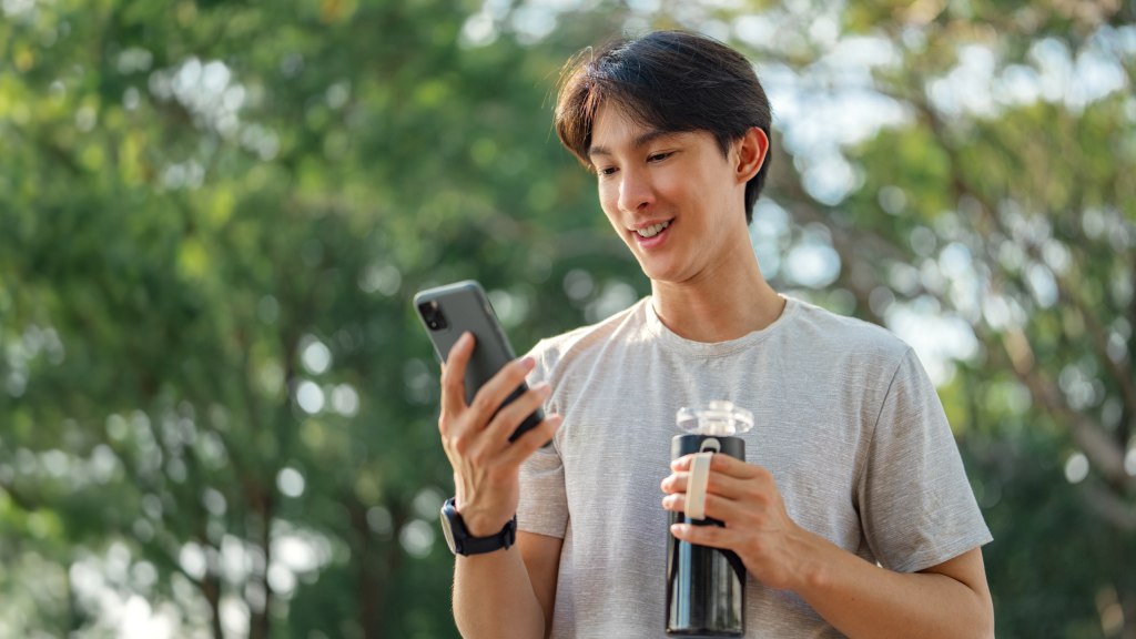 Rapaz jovem com camiseta cinza segurando garrafa d’água em uma mão e celular na outra, sorrindo ao ar livre com árvores ao fundo enquanto usa seus dados móveis fora de casa.