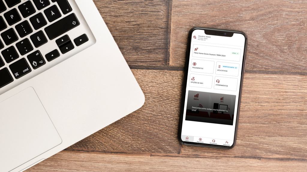 Celular sobre mesa de madeira com o app da Desktop aberto. Ao lado, um notebook é mostrado parcialmente.