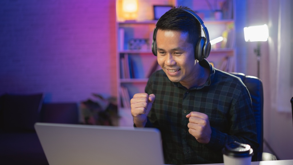 Rapaz gamer com camisa xadrez e headphones pretos, sentado em frente à um notebook gamer, com iluminação em tons de azul, roxo e rosa ao fundo, vibrando durante partida de jogo online.