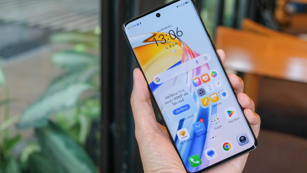 Mão segurando celular Android grande que exibe horário, clima, barra de pesquisa do Google e diversos ícones de aplicativos, representando uma das versões do sistema Android.