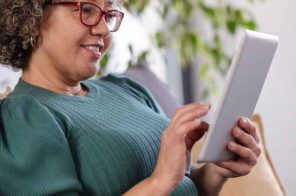 Mulher com blusa verde, calças azuis e óculos de grau com armação vermelha sorrindo sentada no sofá com tablet na mão enquanto traduz páginas da web para ler conteúdos.