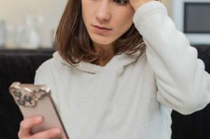 Mulher jovem com blusa branca sentada próximo à cozinha segurando celular iPhone e com a outra mão na cabeça, preocupada por que a rede Wi-Fi não aparece no aparelho.