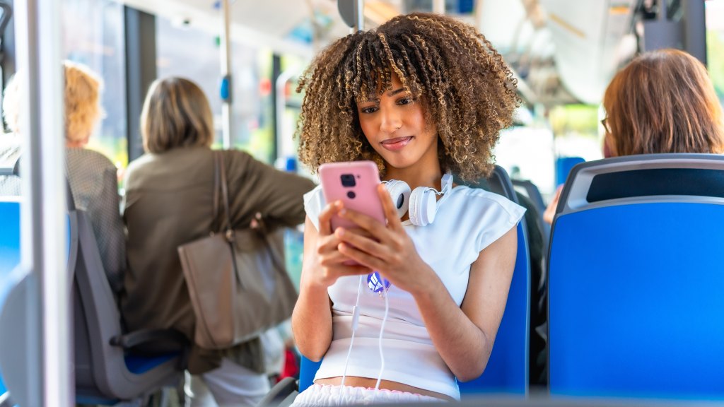 Mulher jovem com blusa branca e headphones brancos no pescoço sentada em transporte público, segurando o celular conectado aos dados móveis, com outras pessoas ao fundo.