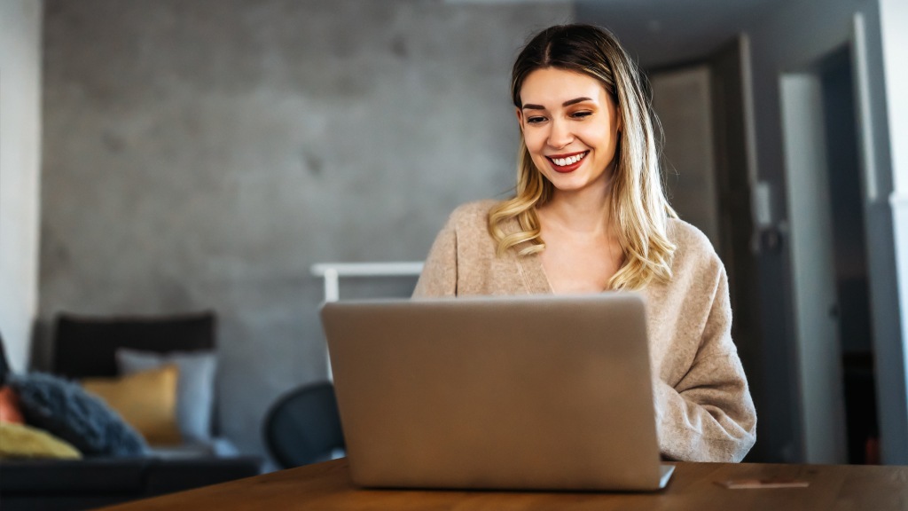 Use o teste de velocidade da Desktop para medir o upload Mulher sorridente com blusa marrom clara sentada à mesa do computador enquanto realiza o teste de velocidade de internet no notebook para conferir a taxa de upload.