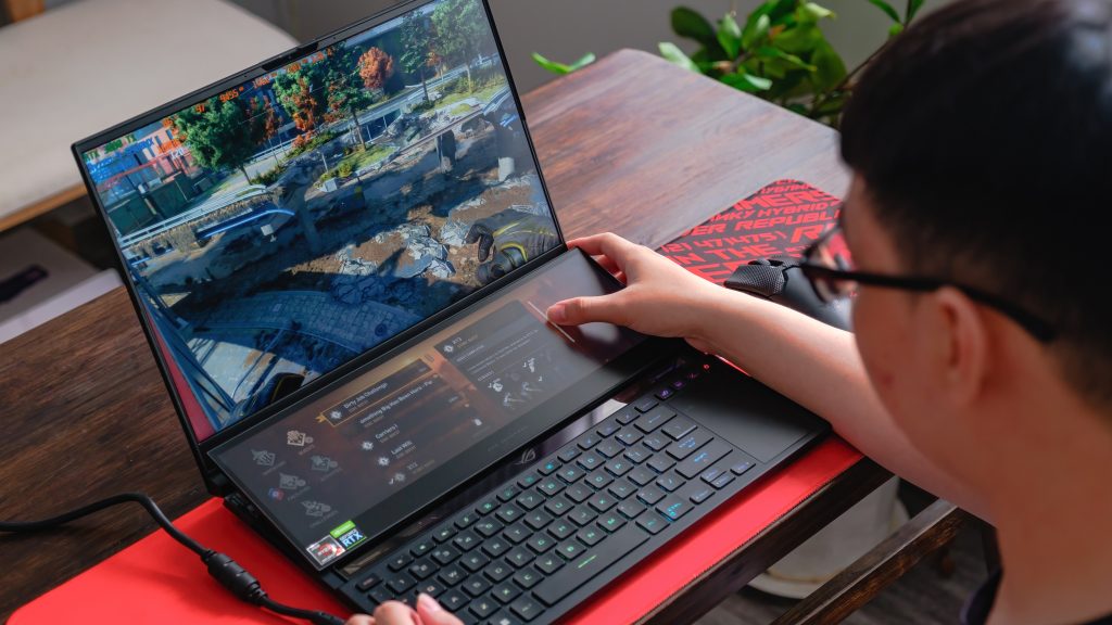 Jovem gamer mostrado parcialmente de perfil, usando óculos, sentado à mesa em frente a um notebook gamer durante partida online.