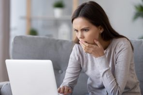 Moça jovem com blusa cinza sentada no sofá, em frente ao notebook, com a mão na boca e expressão de espanto após descobrir que seus dados foram vazados na dark web.