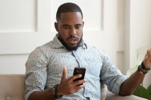 Homem com camisa social azul clara estampada com bolinhas sentado no sofá, segurando o celular em uma mão e com a outra levantada, exibindo expressão de espanto após ter o aparelho hackeado.