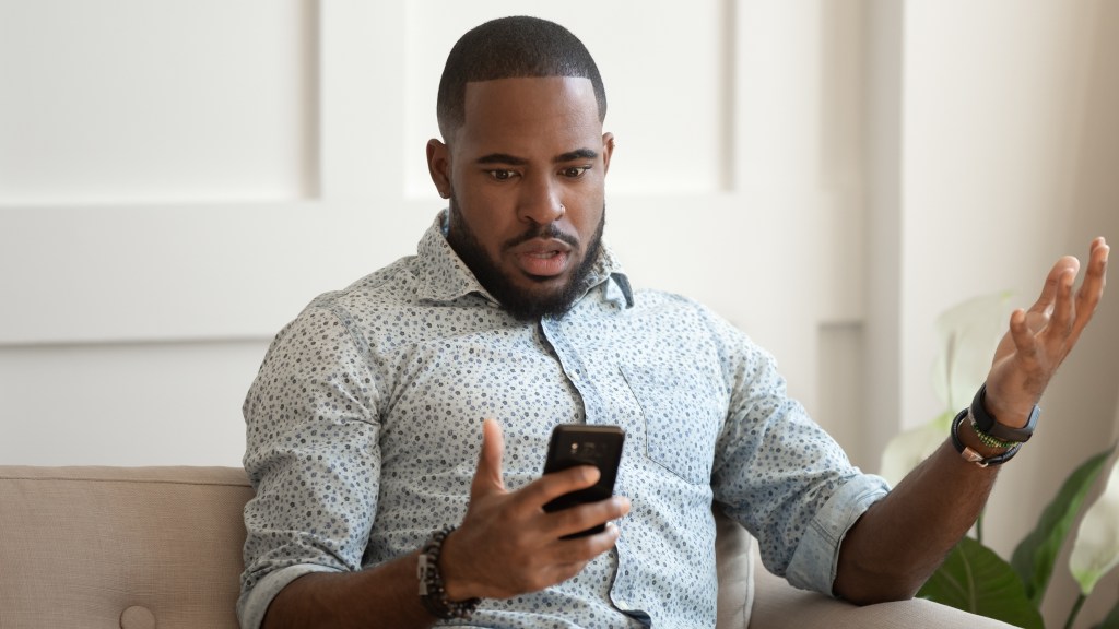 Homem com camisa social azul clara estampada com bolinhas sentado no sofá, segurando o celular em uma mão e com a outra levantada, exibindo expressão de espanto após ter o aparelho hackeado.