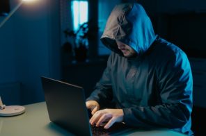 Homem usando blusa com capuz preto, sentado à mesa e navegando em um notebook, representando atividades associadas à dark web.