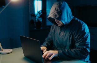 Homem usando blusa com capuz preto, sentado à mesa e navegando em um notebook, representando atividades associadas à dark web.