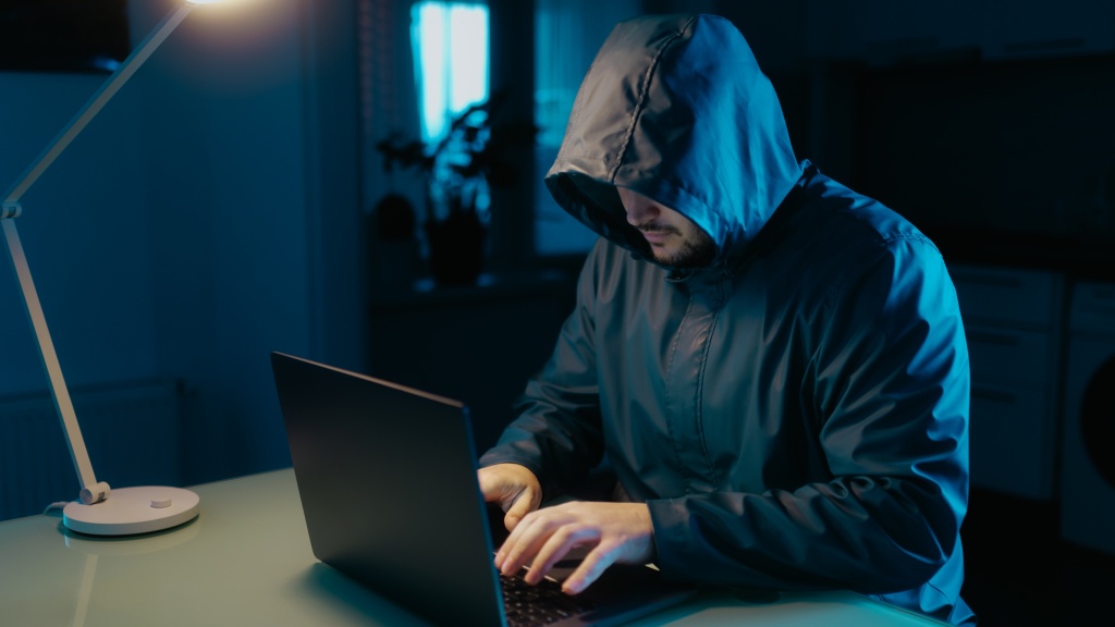 Homem usando blusa com capuz preto, sentado à mesa e navegando em um notebook, representando atividades associadas à dark web.