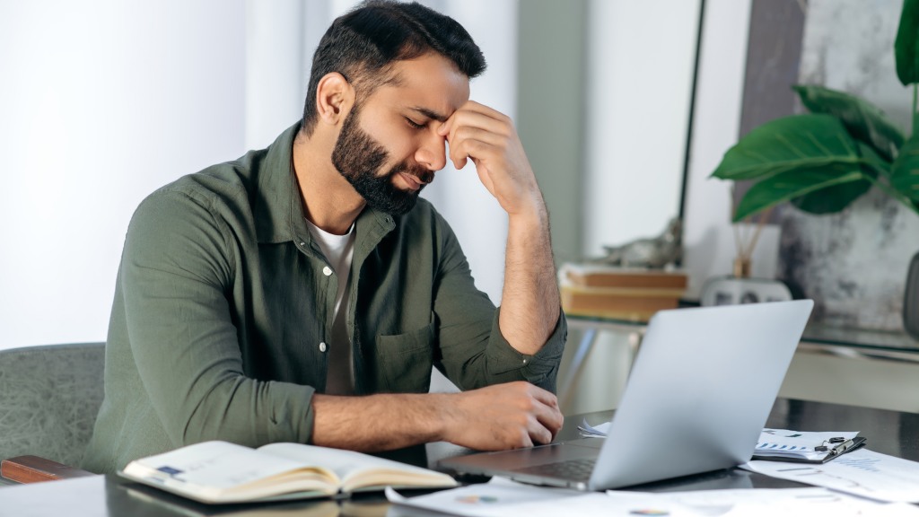 Homem de camisa verde sentado à mesa, em frente ao notebook, com a mão na testa e expressão de frustração após receber spam.