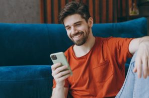 Homem de camiseta vermelha e calça jeans sorrindo, sentado no chão da sala de estar com sofá azul ao fundo, enquanto navega com dados móveis no celular.