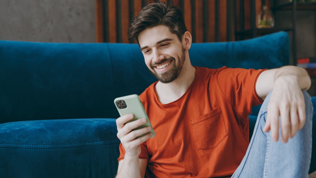 Homem de camiseta vermelha e calça jeans sorrindo, sentado no chão da sala de estar com sofá azul ao fundo, enquanto navega com dados móveis no celular.