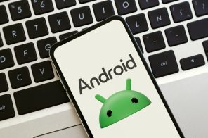 Celular Android sobre teclado de notebook exibindo na tela o rosto do mascote verde do sistema Android e o texto “Android”.