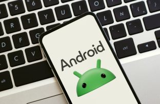 Celular Android sobre teclado de notebook exibindo na tela o rosto do mascote verde do sistema Android e o texto “Android”.