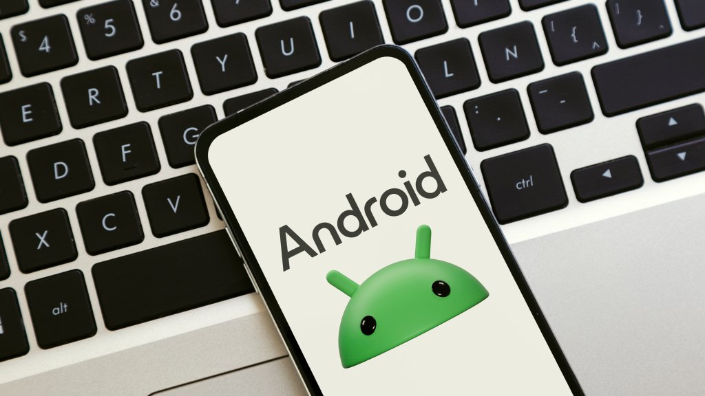 Celular Android sobre teclado de notebook exibindo na tela o rosto do mascote verde do sistema Android e o texto “Android”.