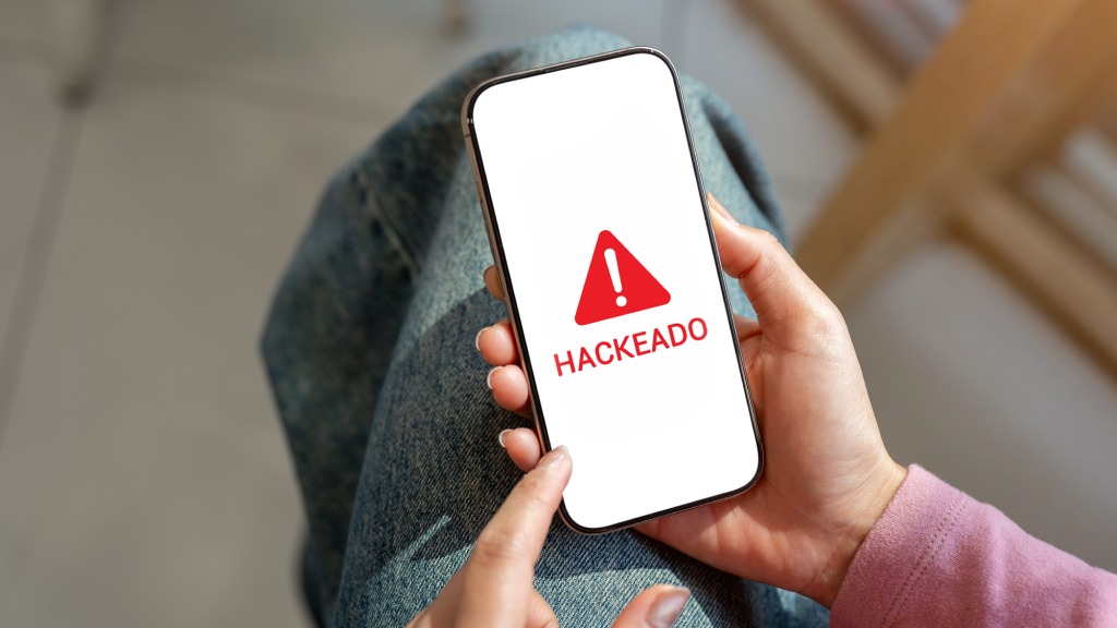 Mão feminina segurando celular, que exibe na tela um ícone de exclamação vermelho e o texto "hackeado", ilustrando dicas para saber se o celular foi hackeado.