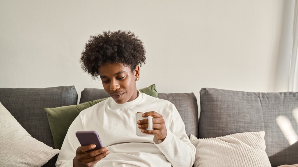 Rapaz com blusa branca sentado em sofá cinza na sala de estar, parede branca ao fundo, segurando caneca branca e celular enquanto aproveita as vantagens do sistema Android.