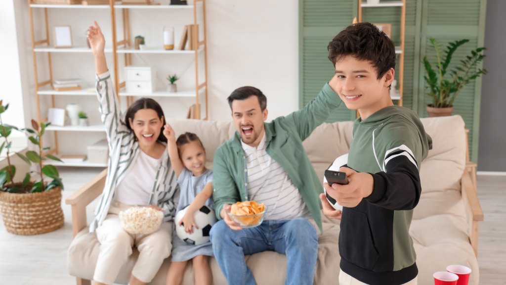 Família com pai, mãe e filha sentados no sofá, com um dos braços erguidos, enquanto outro filtro de pé aparece à frente, apontando o controle para a TV conectada à internet da Desktop, assistindo a conteúdos sem delay.