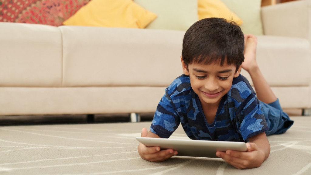 Menino com camiseta camuflada azul deitado de bruços no tapete da sala enquanto assiste a streamings infantis no tablet durante as férias.