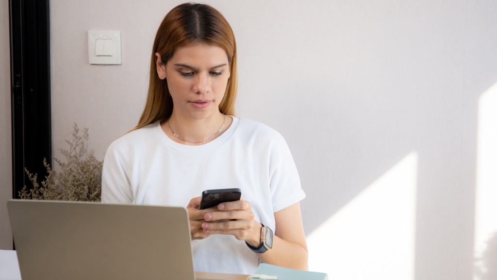 Mulher jovem com camiseta branca sentada à mesa em frente a um notebook enquanto atualiza o software do celular.