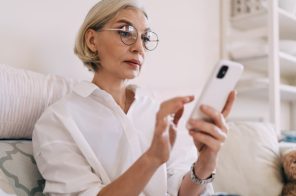 Mulher de meia idade com blusa branca e óculos redondos sentada no sofá, segurando um celular enquanto realiza a atualização do aparelho.