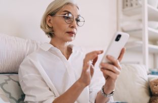 Mulher de meia idade com blusa branca e óculos redondos sentada no sofá, segurando um celular enquanto realiza a atualização do aparelho.