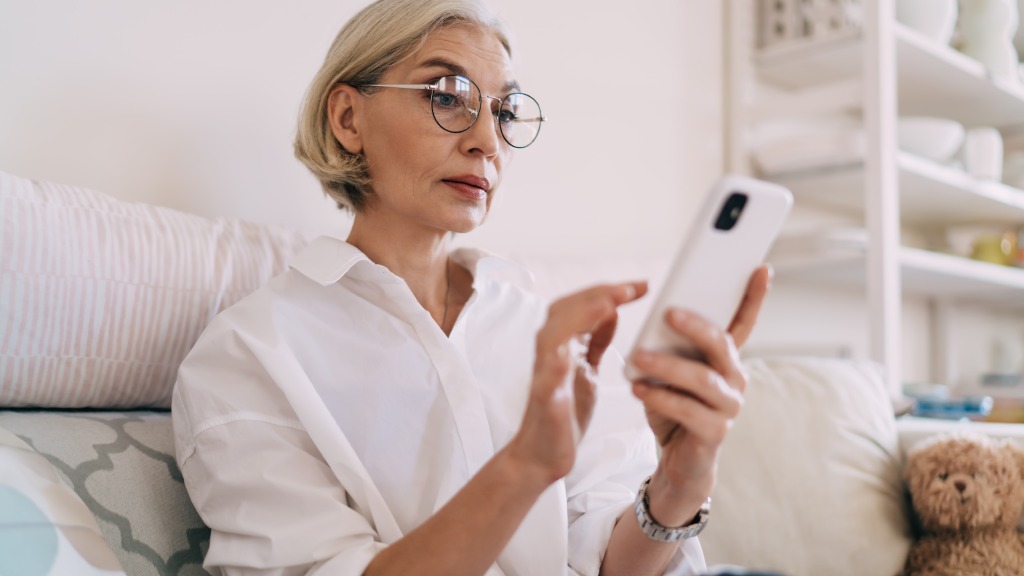 Mulher de meia idade com blusa branca e óculos redondos sentada no sofá, segurando um celular enquanto realiza a atualização do aparelho.