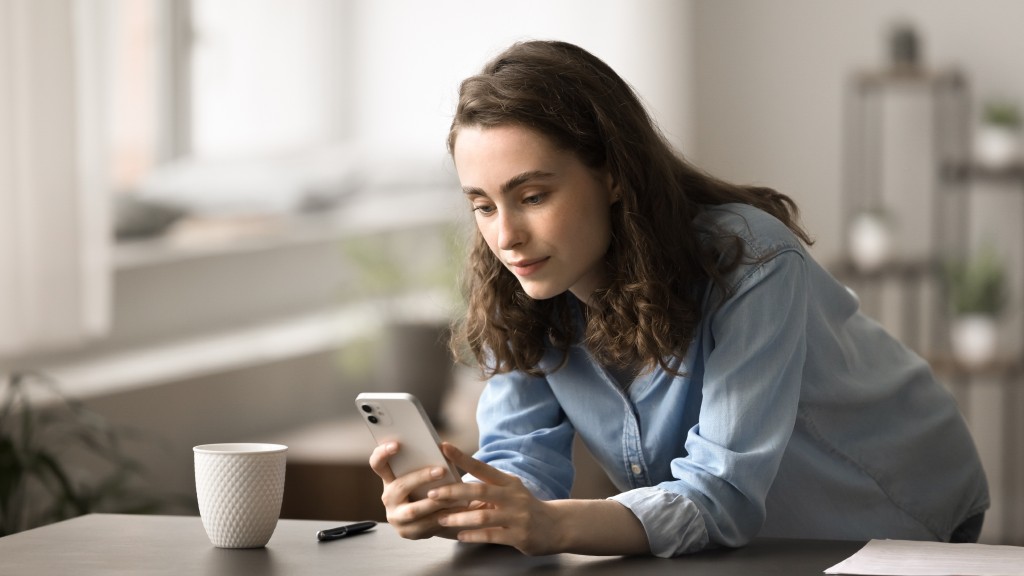 Mulher jovem com camisa jeans azul clara de pé, inclinada com os cotovelos apoiados na mesa, usando o celular concentrada para compartilhar gravações de tela do aparelho.