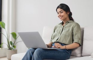 Mulher jovem com camisa verde e calças jeans sentada no sofá da sala, sorrindo, com um notebook cinza no colo, consultando a disponibilidade de internet fibra óptica no seu endereço.