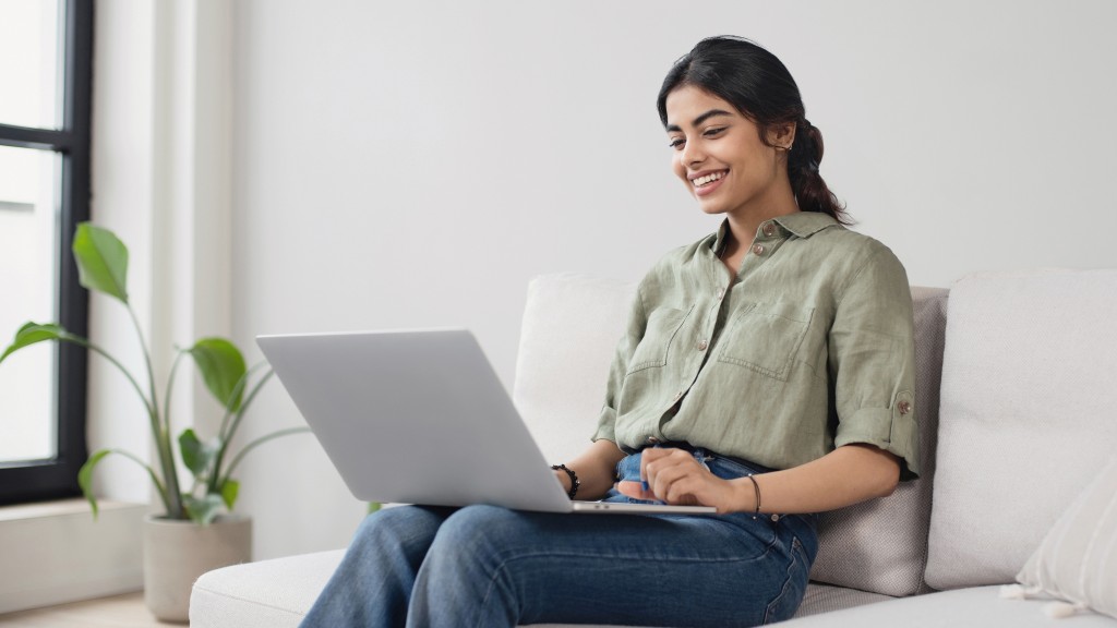 Mulher jovem com camisa verde e calças jeans sentada no sofá da sala, sorrindo, com um notebook cinza no colo, consultando a disponibilidade de internet fibra óptica no seu endereço.