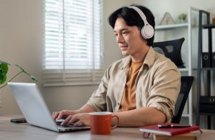 Homem jovem com camisa bege e headphones brancos, sentado à mesa do escritório enquanto converte arquivos no notebook.