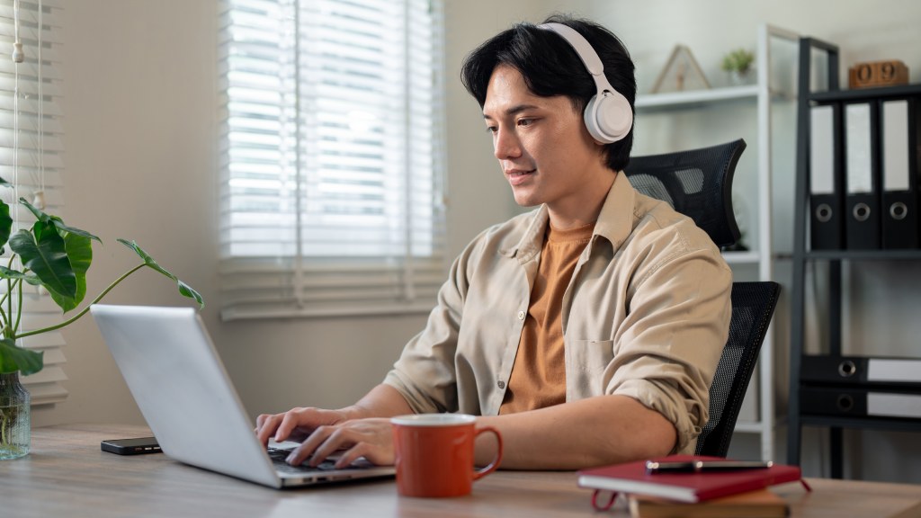 Homem jovem com camisa bege e headphones brancos, sentado à mesa do escritório enquanto converte arquivos no notebook.