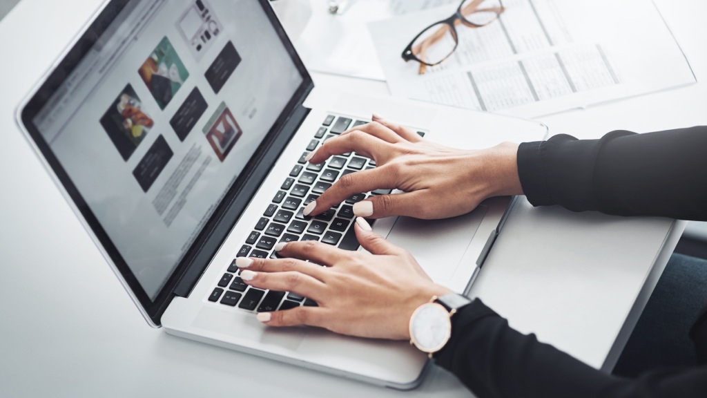 Mãos de uma mulher, com um relógio redondo no pulso esquerdo, sobre o teclado de um notebook que exibe na tela arquivos de imagens sendo convertidos.
