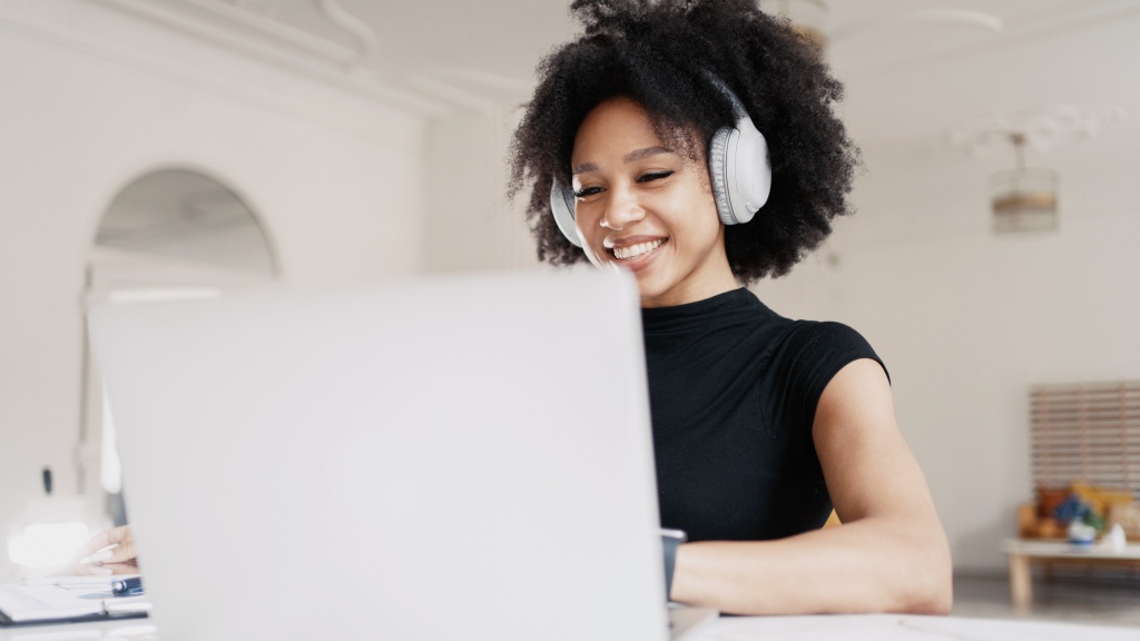 Mulher jovem usando headphones brancos, sentada em frente ao notebook e sorrindo enquanto navega na internet conectada a um roteador mesh.