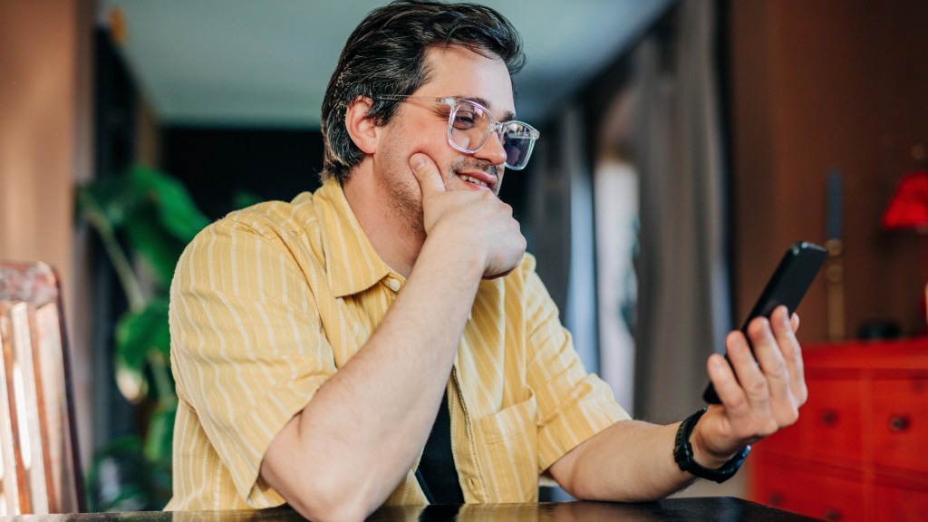 Homem com camisa amarela com listras brancas e óculos de armação transparente, sentado à mesa de casa, com uma mão no queixo e a outra segurando o celular conectado ao Plano Controle da Desktop.
