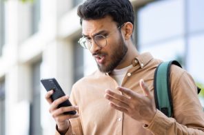 Homem jovem com camisa marrom clara, óculos redondos e mochila nas costas, de pé ao ar livre, com expressão de indignação ao olhar para o celular com memória cheia, representando como liberar espaço no aparelho.