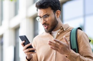 Homem jovem com camisa marrom clara, óculos redondos e mochila nas costas, de pé ao ar livre, com expressão de indignação ao olhar para o celular com memória cheia, representando como liberar espaço no aparelho.