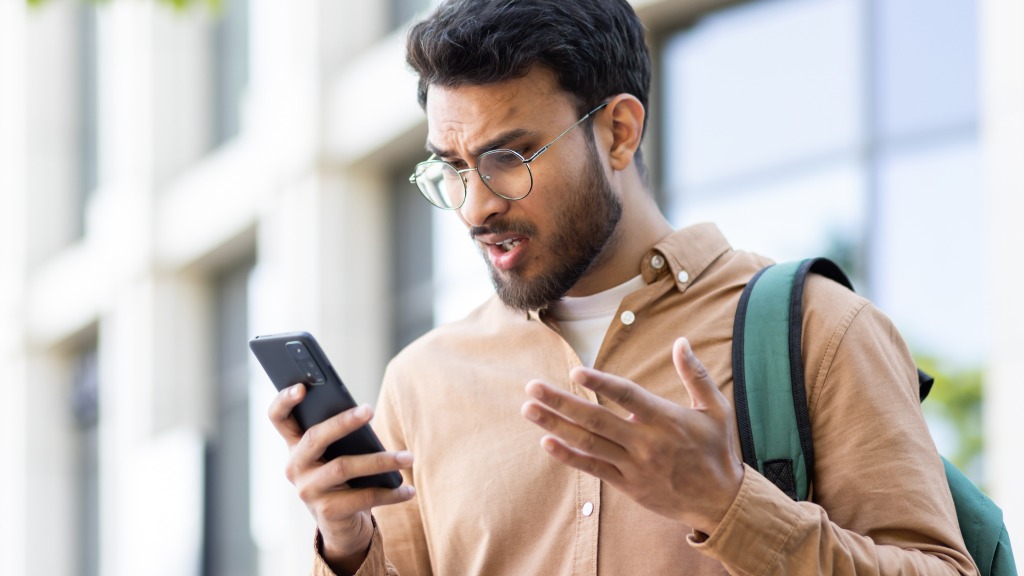 Homem jovem com camisa marrom clara, óculos redondos e mochila nas costas, de pé ao ar livre, com expressão de indignação ao olhar para o celular com memória cheia, representando como liberar espaço no aparelho.