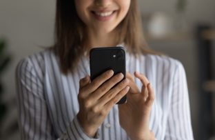 Mulher jovem com camisa branca de listras azuis, desfocada, sorrindo enquanto segura o celular e ativa o aplicativo Celular Seguro.