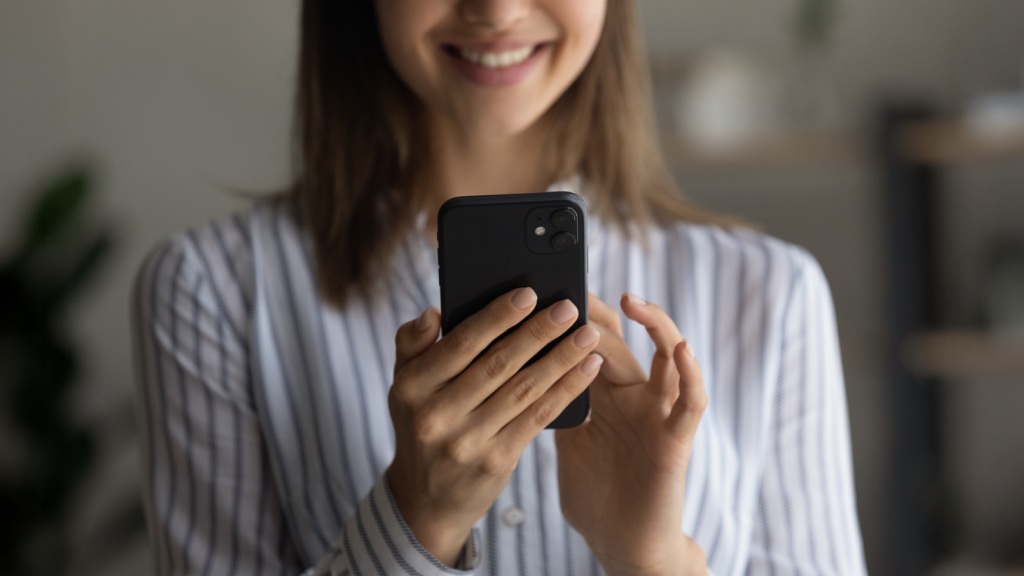 Mulher jovem com camisa branca de listras azuis, desfocada, sorrindo enquanto segura o celular e ativa o aplicativo Celular Seguro.