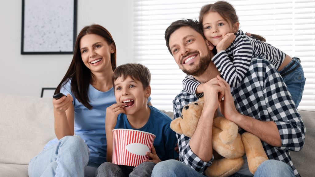 Família com esposa, marido e duas crianças — um menino e uma menina — sentados no sofá da sala e sorrindo. A mãe aponta o controle para a TV, o filho segura um balde de pipoca e o pai tem uma pelúcia no colo, enquanto aproveitam os benefícios após trocar de internet.