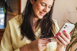 Mulher jovem com blusa amarela sentada em uma cadeira em casa, sorrindo enquanto segura o celular e um cartão para pagar a fatura Desktop pelo aplicativo.