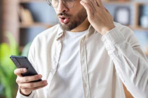 Homem com camisa bege e camiseta azul, de pé em casa, com uma das mãos na lateral da cabeça e a outra segurando o celular, com expressão de espanto ao identificar que o aparelho tem vírus.