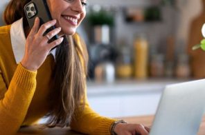 Mulher com blusa amarela, sorrindo, apoiada no balcão da cozinha enquanto fala ao telefone e digita no notebook, representando uma cliente entrando em contato pelo número da Desktop.