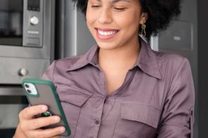 Mulher com blusa roxo escuro, sorrindo, de pé e apoiada no balcão da cozinha, segurando uma xícara amarela e um iPhone enquanto fala com o WhatsApp da Desktop.