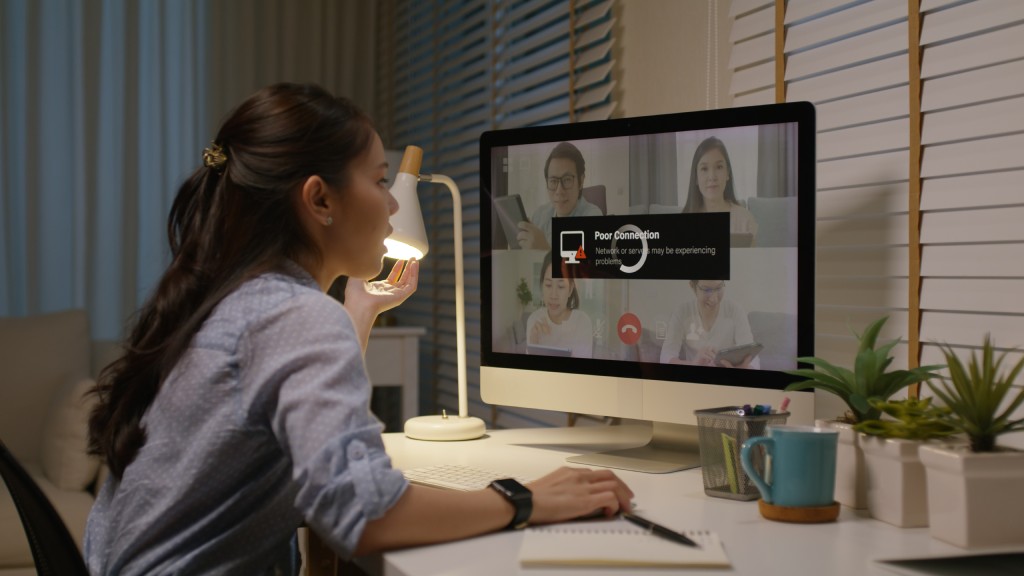 Mulher jovem com camisa azul clara sentada na escrivaninha do quarto, em frente ao computador de mesa, cujo monitor exibe quatro pessoas em uma videochamada com delay e a notificação “Poor connection” (“Conexão ruim”).