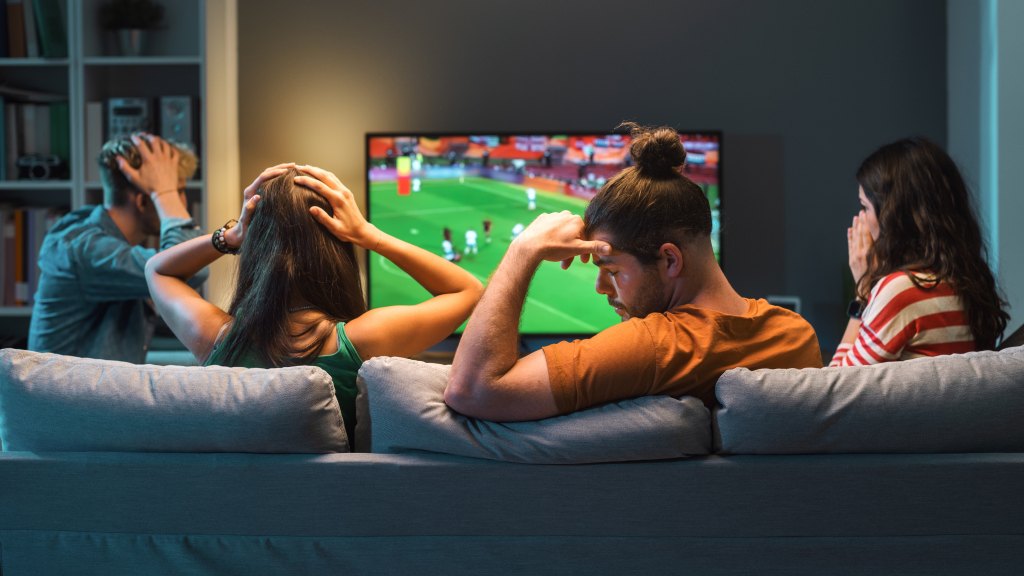 Quatro pessoas vistas de trás — dois homens e duas mulheres — sentadas no sofá assistindo futebol na TV, com as mãos na cabeça ou no rosto, demonstrando frustração com o delay na internet durante a transmissão ao vivo.